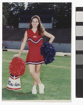 A color photograph of a young, smiling White woman in a red cheerleader uniform, holding pom-poms and leaning on a megaphone on a grassy sports field.