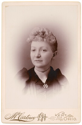 A sepia-toned studio portrait photograph of a White woman with curly, light colored hair in an updo, wearing a dark top closed with a brooch at the neck.