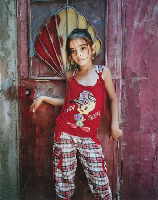 A color portrait photograph of a young medium-skinned girl wearing a red sleeveless shirt and red plaid pants standing in front of a door.