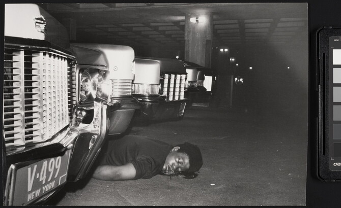 A black-and-white photograph of the body of a Black person on their stomach with a blood stain around their head between cars in a parking garage.