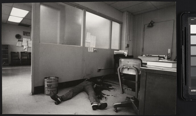 A black-and-white photograph of an office with a bloody body on the floor, feet toward the camera, the top part of the body obscured by dark stains on it, the floor, and the wall.