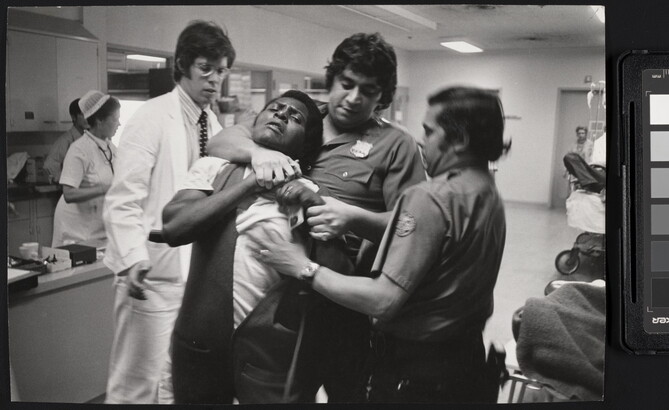 A black-and-white photograph of a Black man being restrained and choked by two police officers with medium to dark skin in a hospital setting.