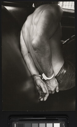 A black-and-white photograph of the bare back and handcuffed hands of a White man in the back seat of a police car.