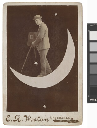 A black-and-white photograph of a White man wearing a bowler hat and suit holding an old-fashioned camera on a tripod while standing on a crescent moon.