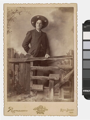 A sepia-toned photograph of a White man wearing a large sombrero-like hat standing behind a split-rail fence that is part of a studio set.