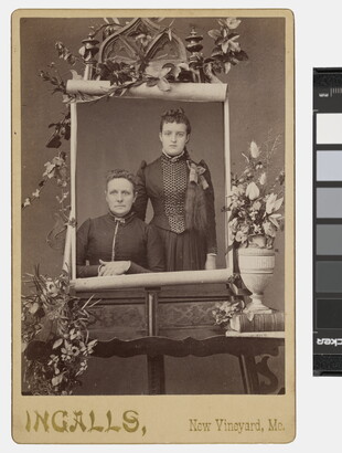 A sepia-toned studio portrait photograph of two White women on a massive paper scroll mounted on an easel and surrounded by flowers.