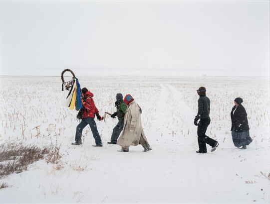 A color photograph of a group of people crossing a vast, snowy landscape.