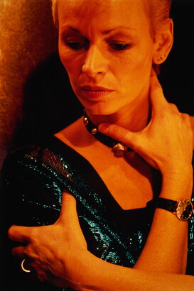 A color portrait photograph of a White woman, from forehead to upper arms, wearing a shimmering blue top, one arm wrapped around an upper arm, the other reaching up to her neck.