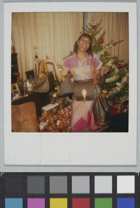 A color Polaroid photograph of a Black woman gleefully showing off several designer purses in front of a Christmas tree.