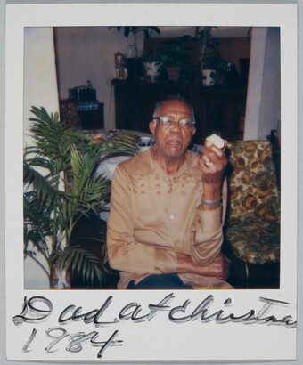 A color Polaroid photograph of a seated, middle-aged Black man in glasses and a light brown shirt holding an apple in one hand; handwritten text at bottom reads: &quot;Dad at Christma[s] 1984.&quot;