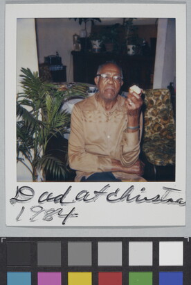 A color Polaroid photograph of a seated, middle-aged Black man in glasses and a light brown shirt holding an apple in one hand; handwritten text at bottom reads: &quot;Dad at Christma[s] 1984.&quot;