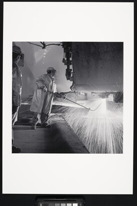 A black-and-white photograph of an industrial worker, wearing safety helmet, coat, and gloves holding a rod under a machine emitting sparks.