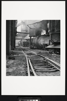 A black-and-white photograph of railroad track  between industrial buildings and  machinery, some emitting steam or smoke.