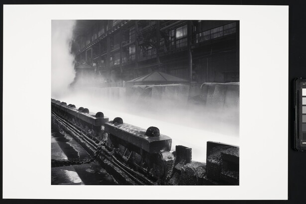 A black-and-white photograph of industrial machinery in front of steam or smoke rising near a multi-story industrial building.