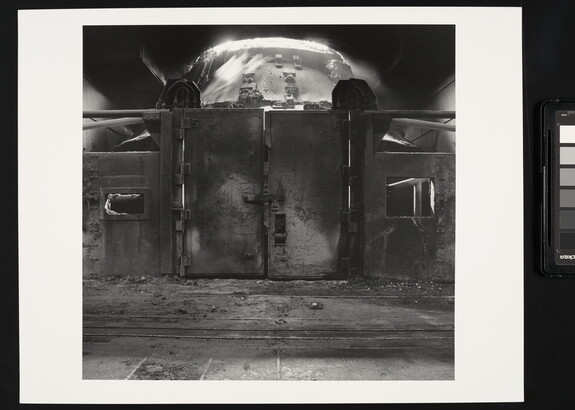 A black-and-white photograph of a metal wall with large doors behind which is an industrial furnace.
