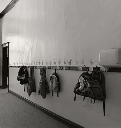 A black-and-white photograph of a hallway with kids&#039; backpacks and jackets hanging on hooks.
