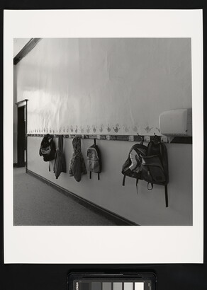A black-and-white photograph of a hallway with kids&#039; backpacks and jackets hanging on hooks.