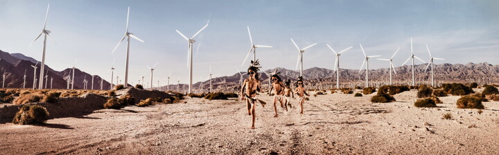 A color photograph of a group of Indigenous children in traditional dress running through a desert landscape covered with wind turbines.