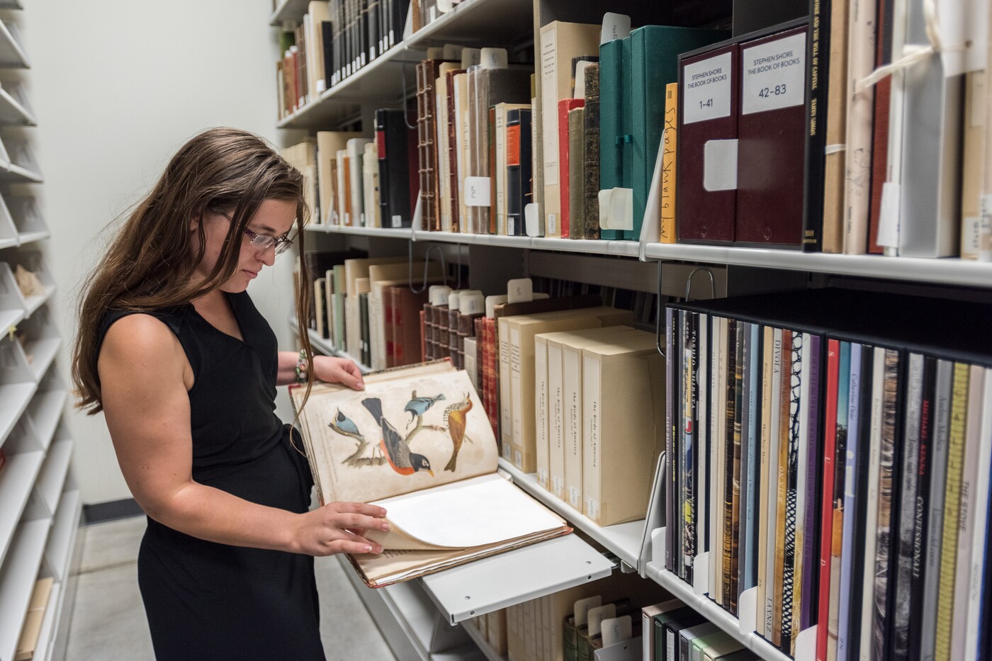 Shelves with archival materials are on the right; on the left a woman has a book open and is reviewing it.