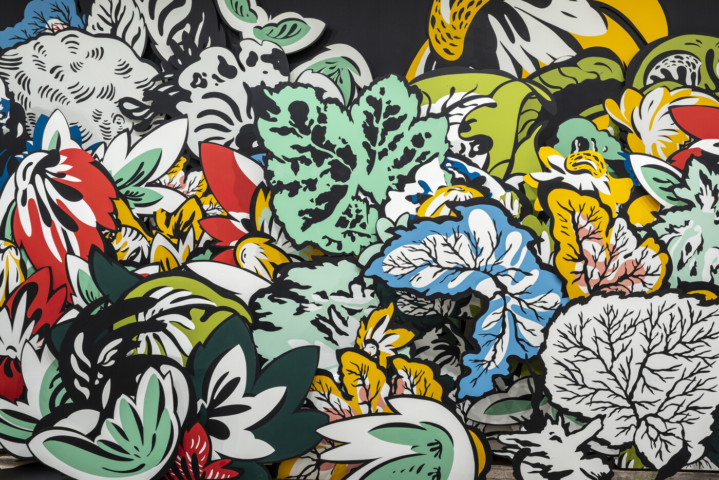 A detail of an installation of large wood cut-outs of flowers and leaves, some brightly-colored, against a black wall with shadows projected on the floor in a Carter gallery. 