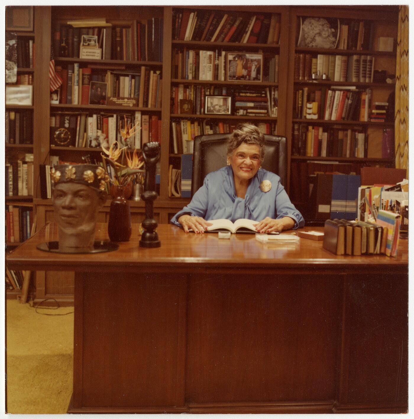 A color photo of an older woman sitting at a desk in a room lined with floor-to-ceiling bookshelves.