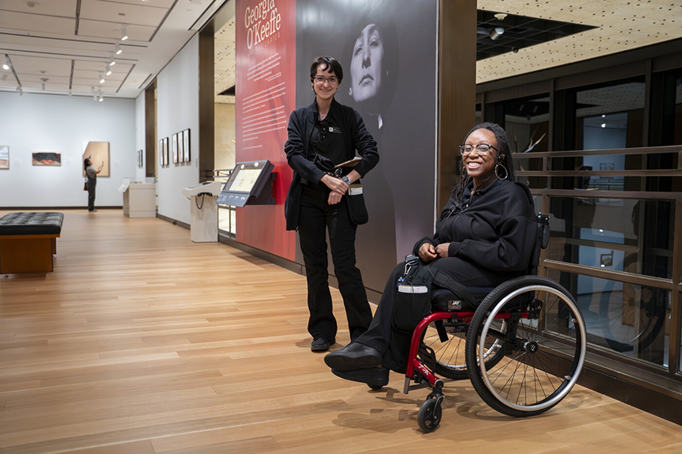 A smiling White woman with short dark hair and a smiling Black woman with long hair seated in a wheelchair, both wearing black, in a Carter gallery.