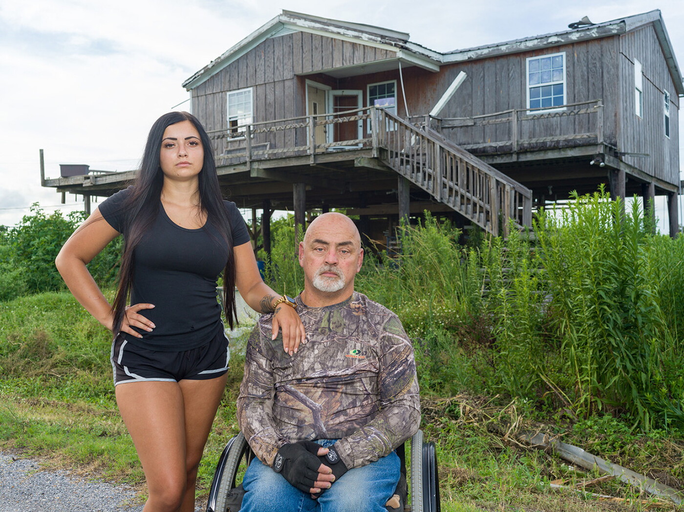 Two people outside of a house on stilts: a young woman with long dark hair stands next to a middle-aged man seated in a wheelchair.