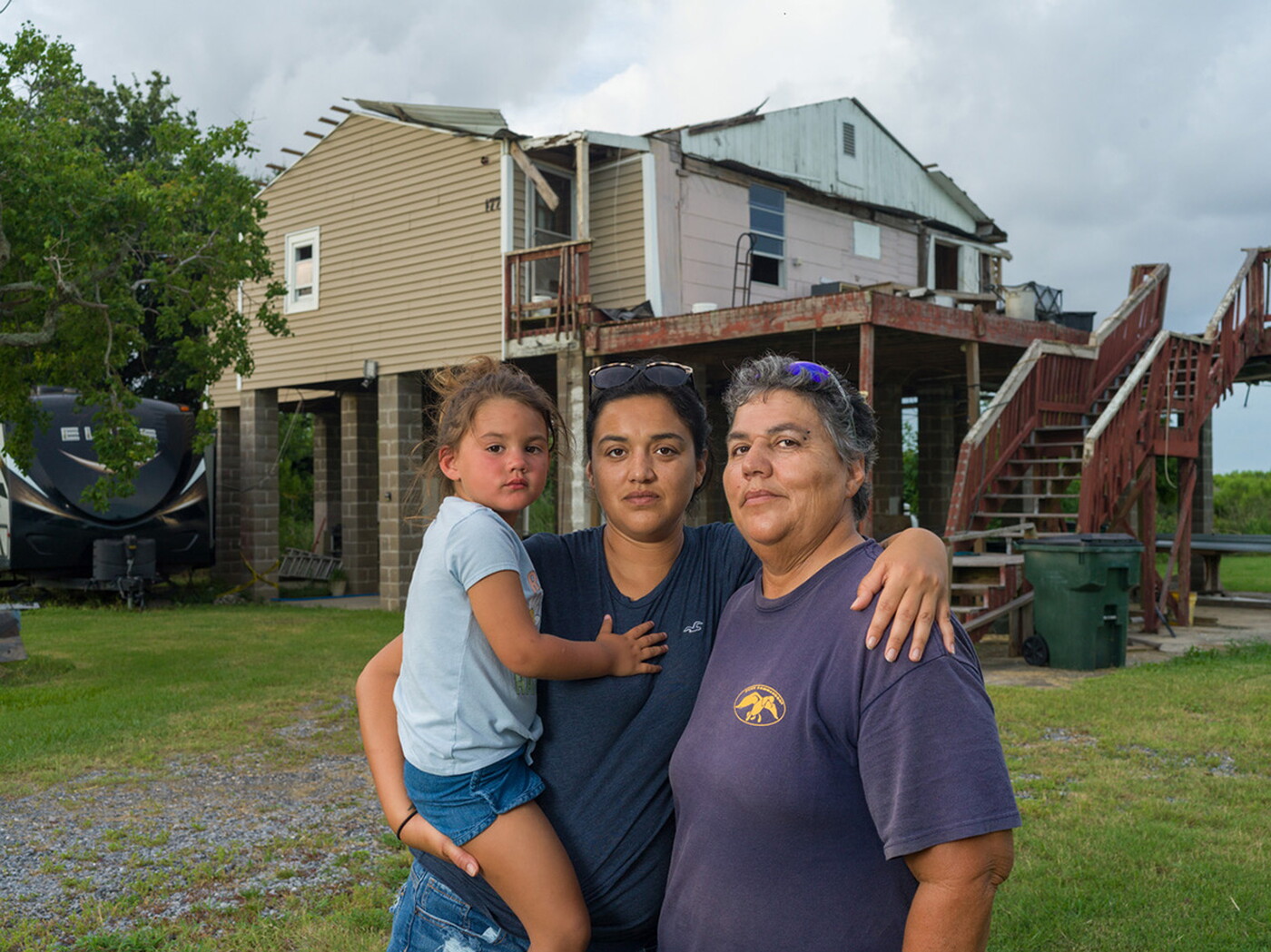 A color photograph of three people in front of a house on stilts: a younger woman holds a child on her hip and has her arm around an older woman.