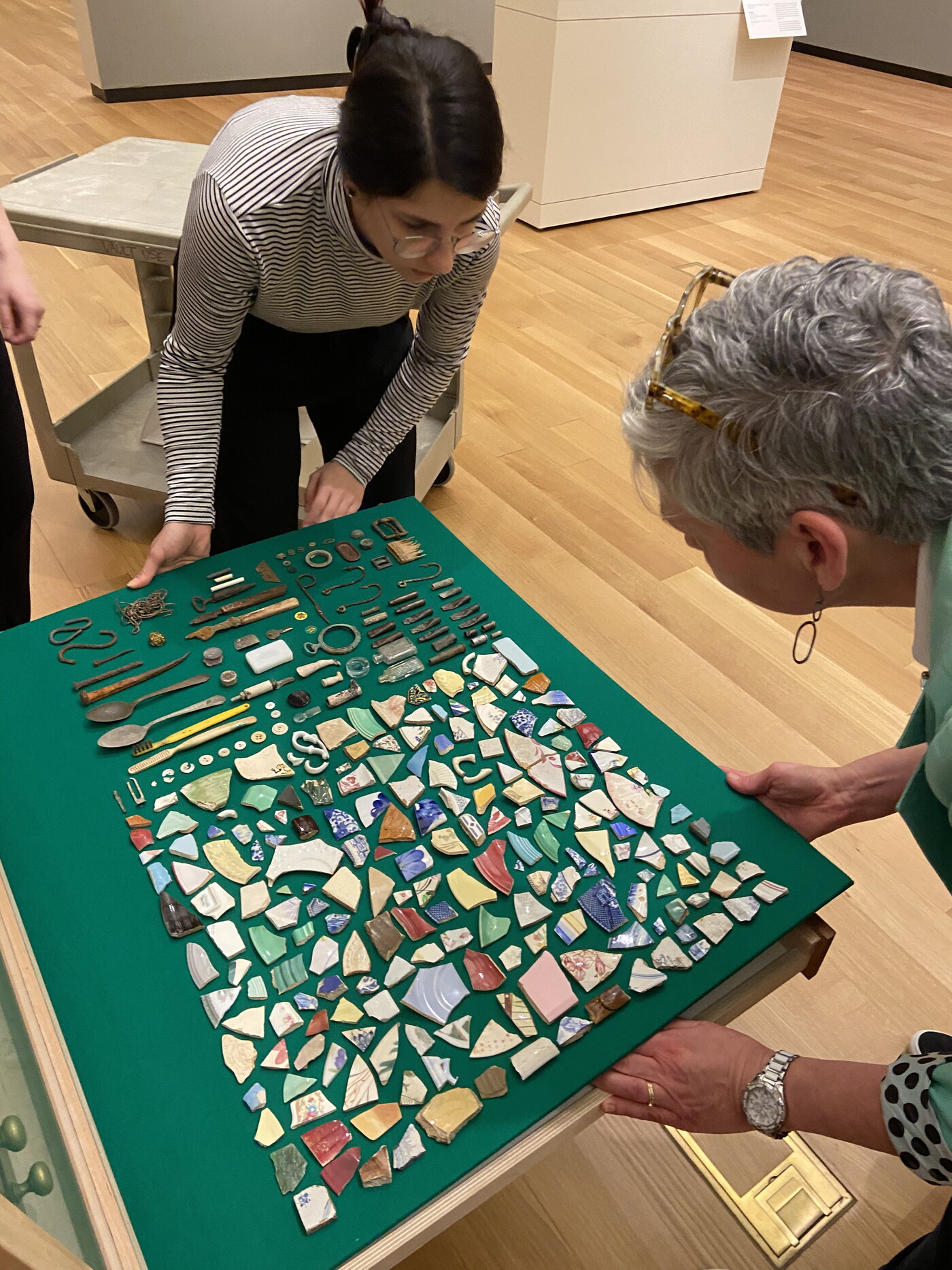 Two women place a green board covered with a variety of objects into a drawer.