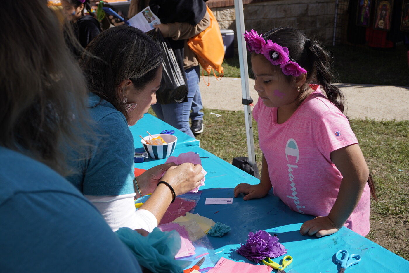 A woman leans over a table to show a medium-skinned child how to make an artwork.