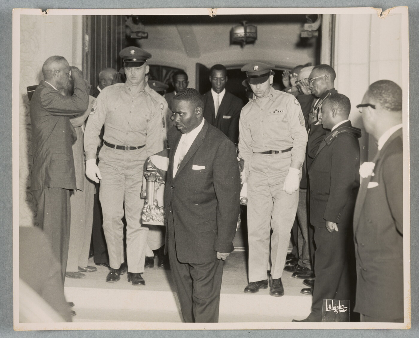 A black-and-white photograph of pallbearers, some in military uniforms, carrying a flag-draped casket from a church as bystanders salute.