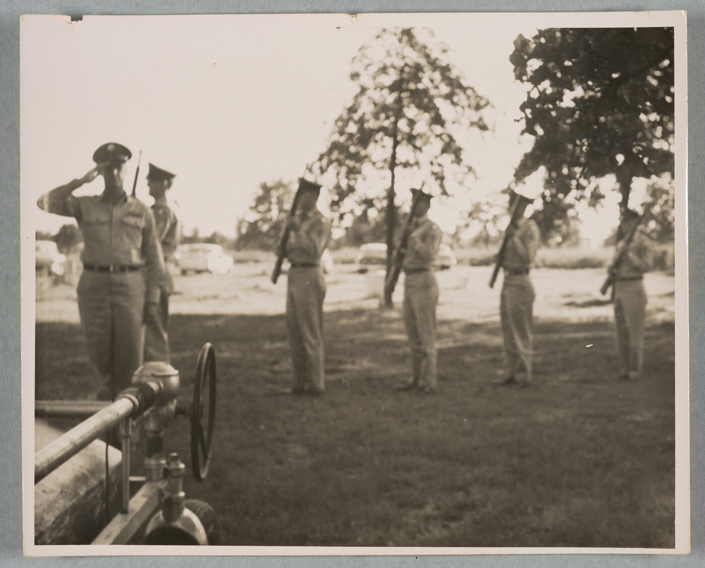 A slightly out-of-focus black-and-white photograph of a line of people in military uniforms, each holding a rifle, as one soldier salutes; tall trees and cars are in the background.
