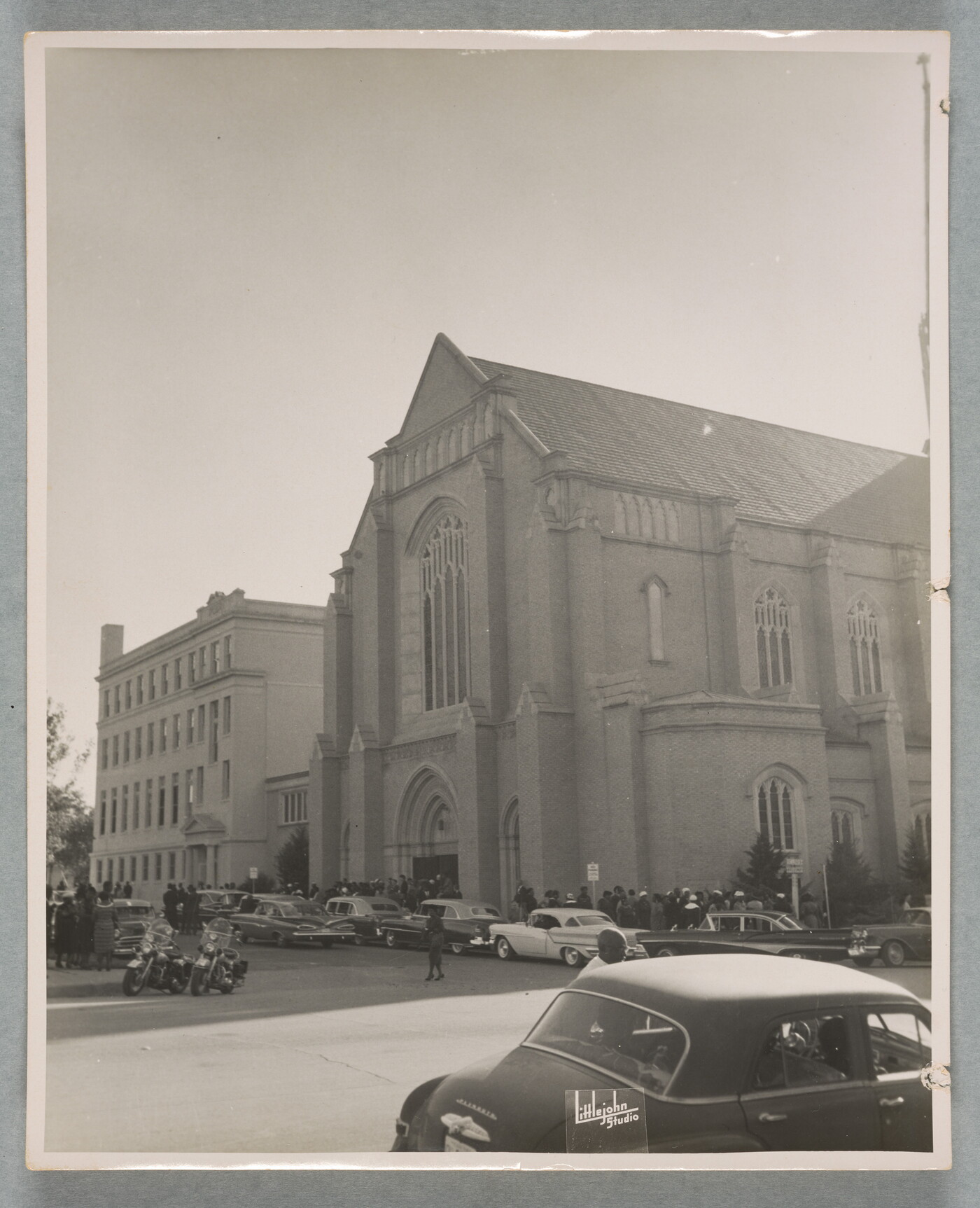 A black-and-white photograph of a large stone church, crowds lined up outside of it and many cars lining the street in front of the church.