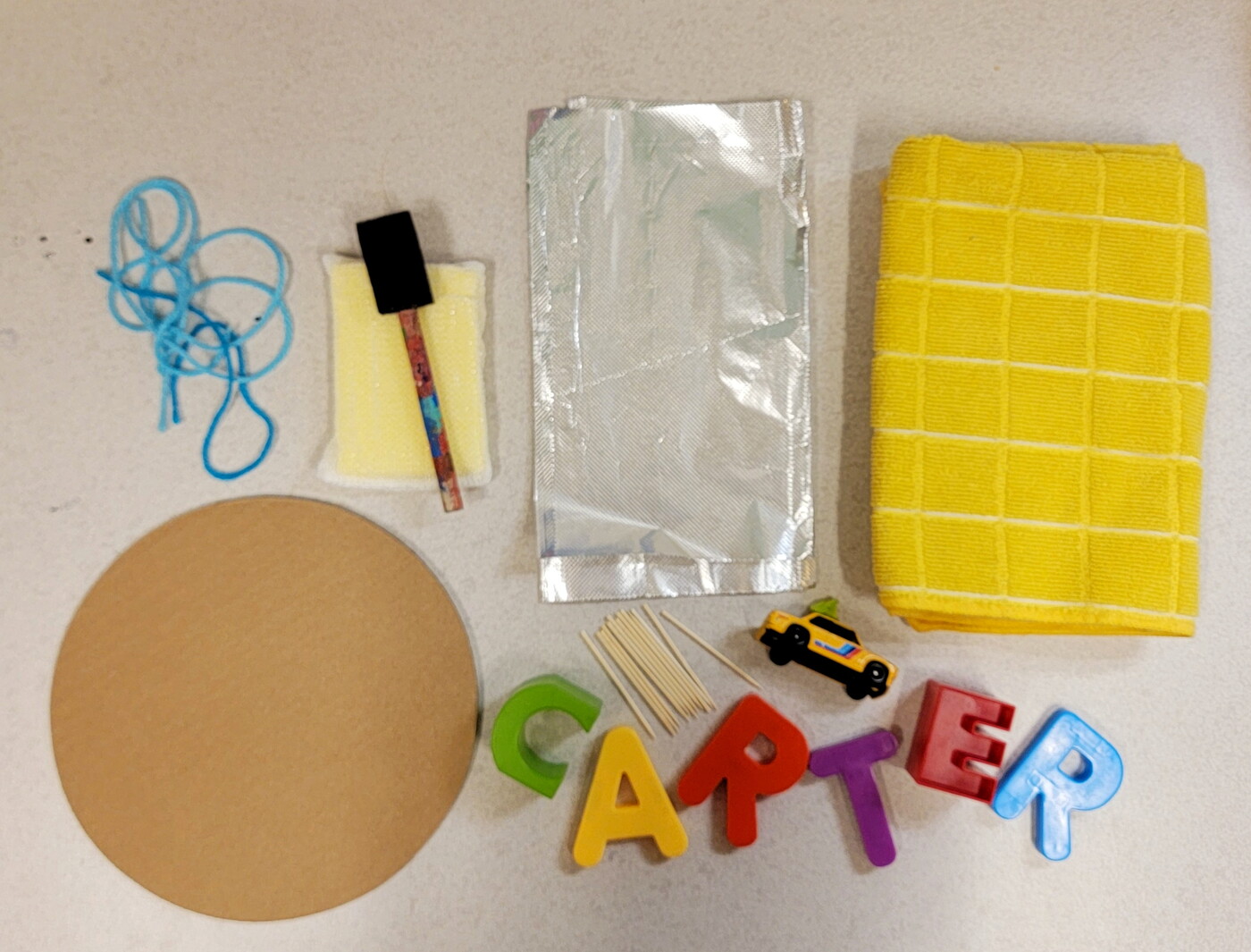 Art supplies layed out on a table: string, cardboard, paint brushes, a sponge, foil, towel, magnetic letters.
