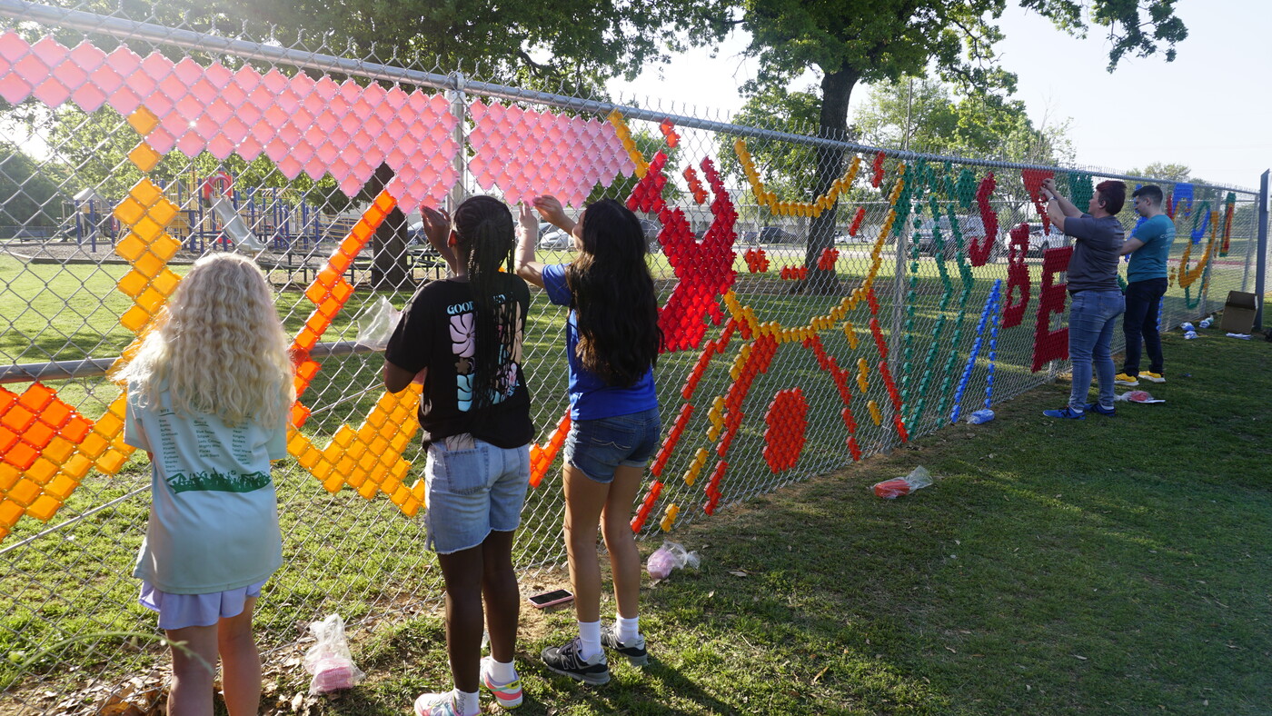 Children and adults create colorful patterns in a chainlink fence using colored plastic pieces.