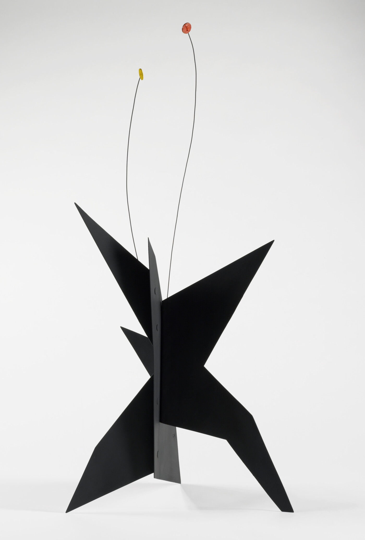 A black, metal, star-like shaped sculpture with two wires rising from the top, one capped with a yellow circle and the other with an orange circle.