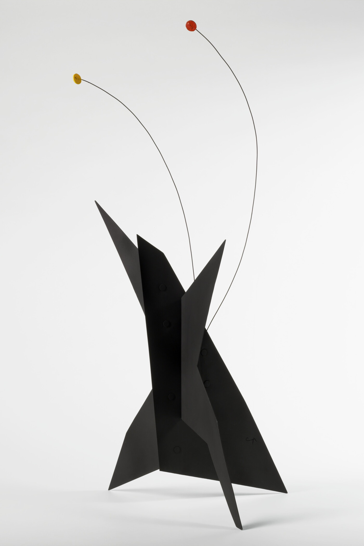 A black, metal, star-like shaped sculpture with two wires rising from the top, one capped with a yellow circle and the other with an orange circle.