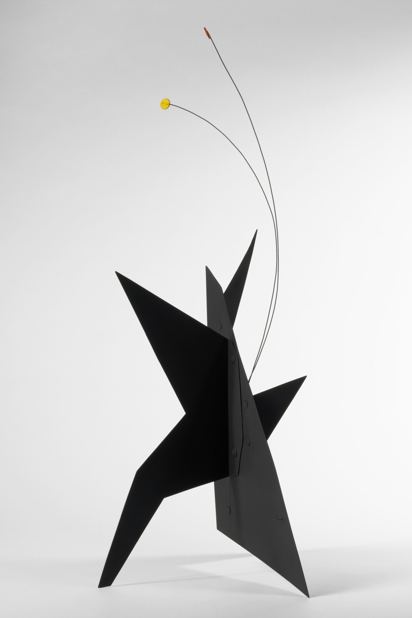 A black, metal, star-like shaped sculpture with two wires rising from the top, one capped with a yellow circle and the other with an orange circle.