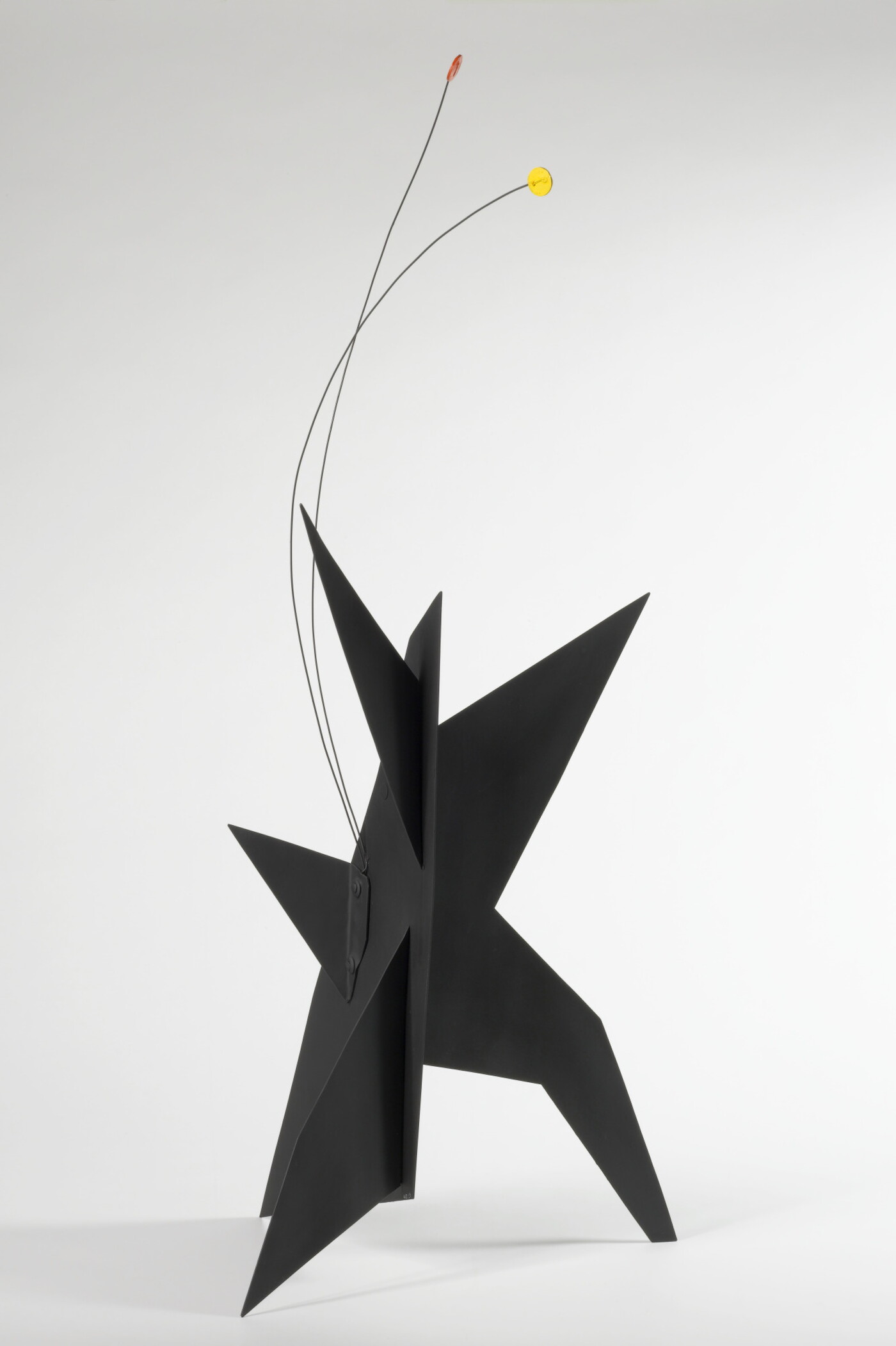 A black, metal, star-like shaped sculpture with two wires rising from the top, one capped with a yellow circle and the other with an orange circle.