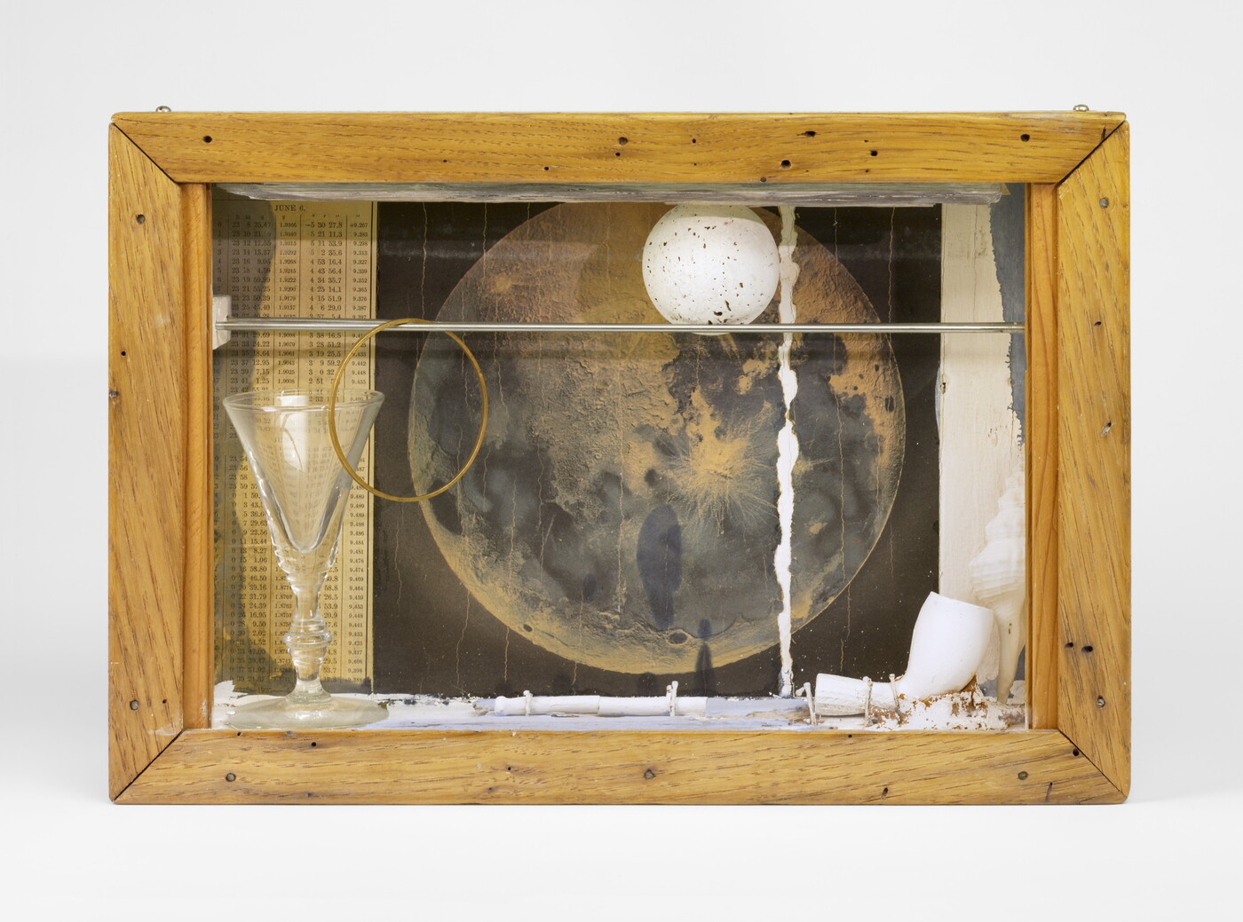 A wood shadowbox containing a pipe, a white ball with gray speckles, a picture of the moon, and a glass goblet.