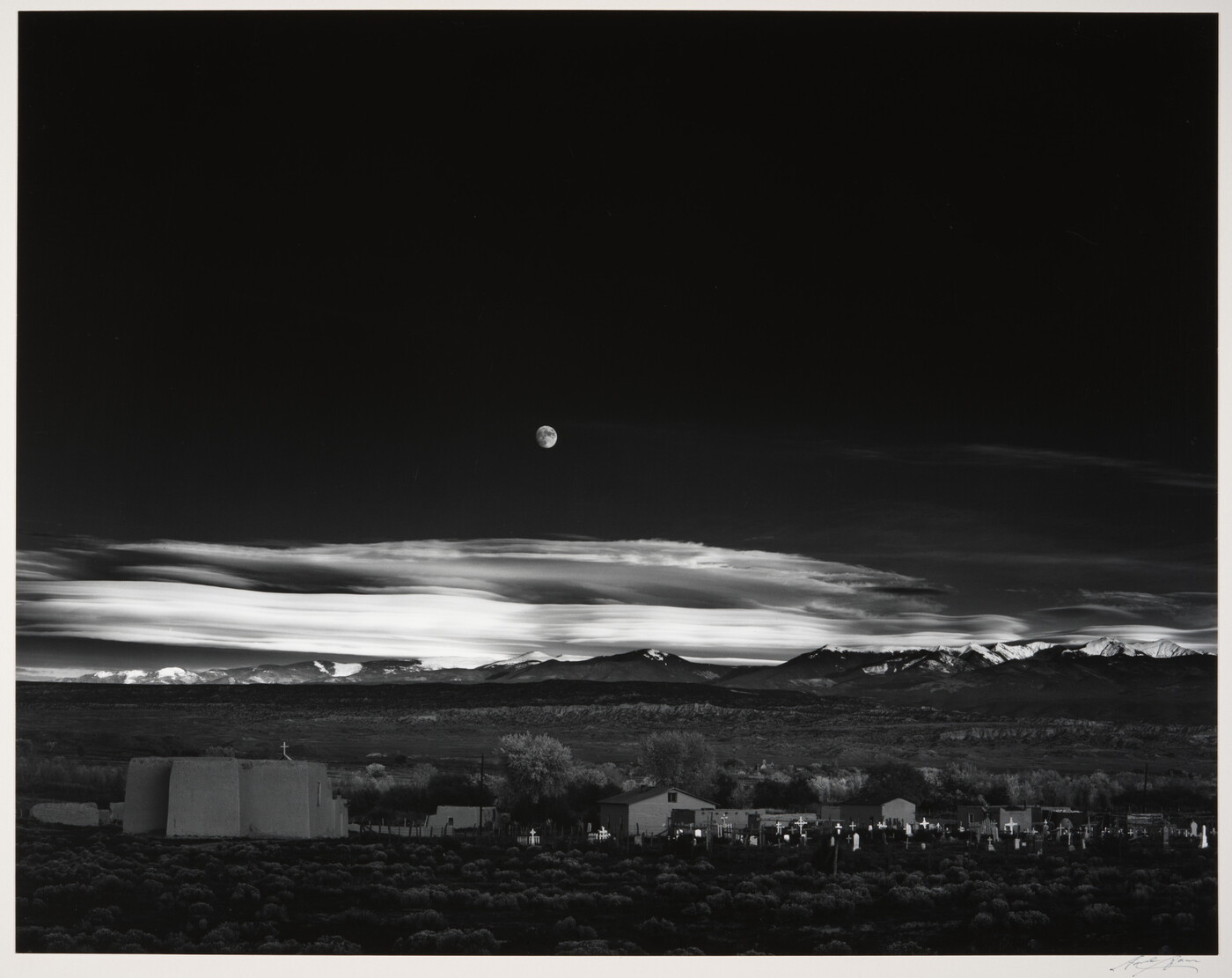 A black-and-white landscape photograph of a town with mountains in the background and the moon rising above the clouds in the night sky.