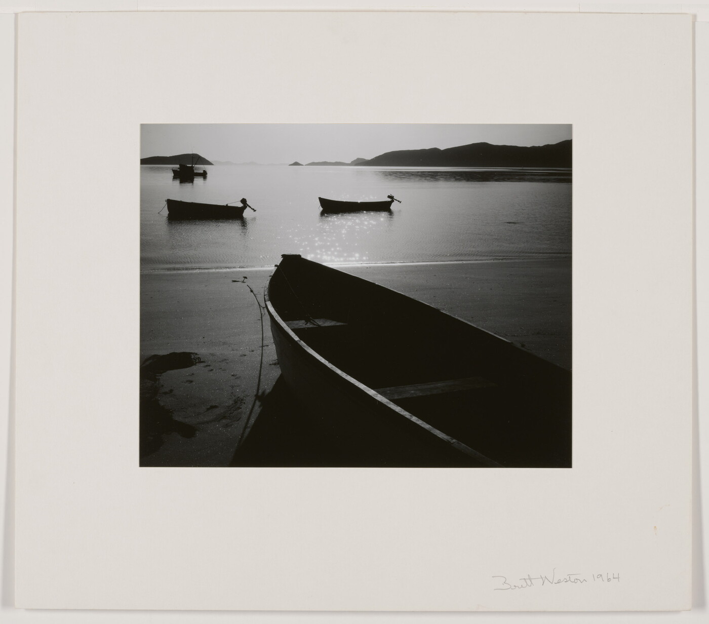 A black-and-white photograph of a rowboat on a sandy beach and three more out on still water with hills in the distance.