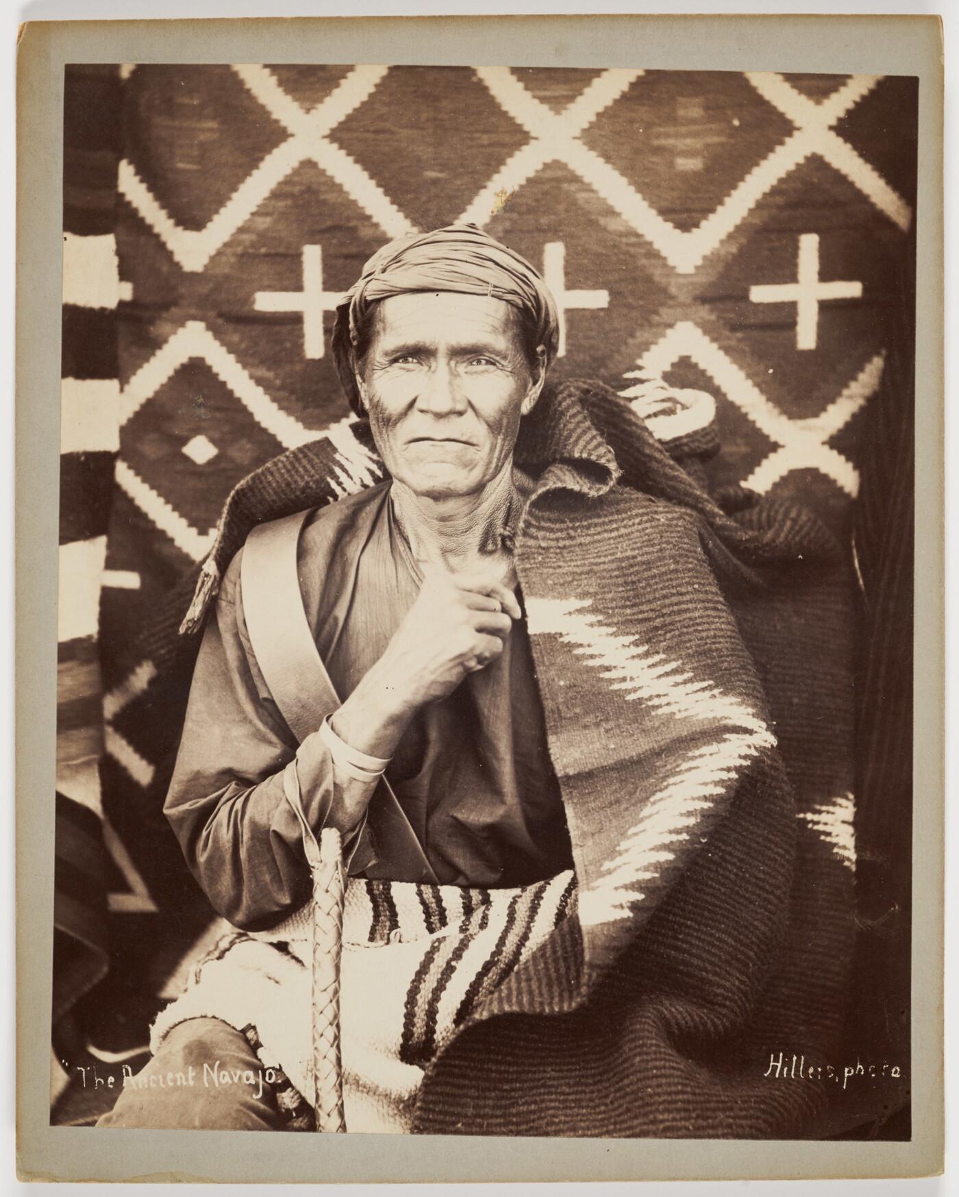 A sepia-toned photograph of an Indigenous person wearing a cloth turban, seated in front of woven blankets with another blanket wrapped around their shoulders.