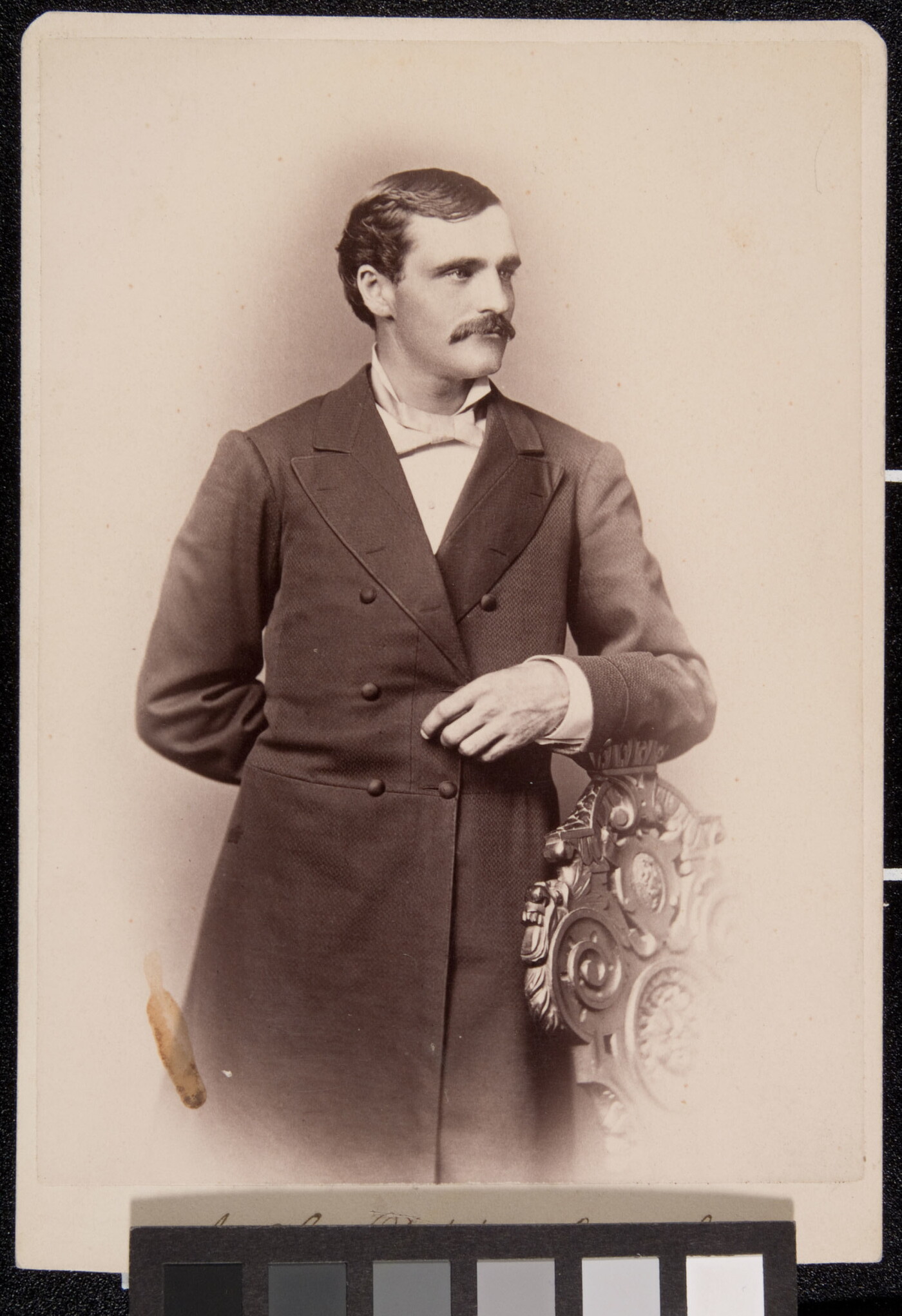A sepia-toned portrait of a White man wearing a long frock coat standing posed with his arm on the back of a chair.