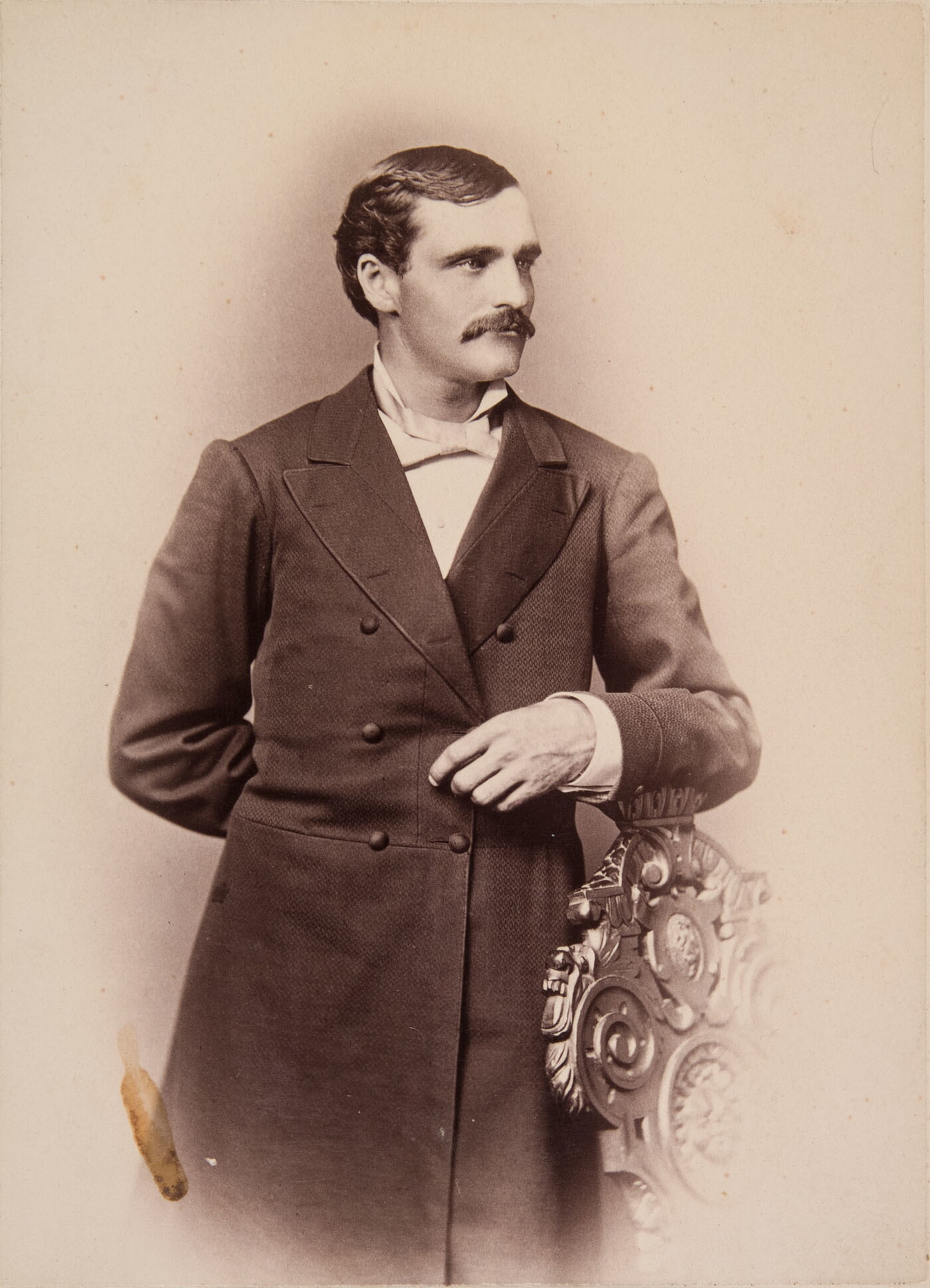 A sepia-toned portrait of a White man wearing a long frock coat standing posed with his arm on the back of a chair.