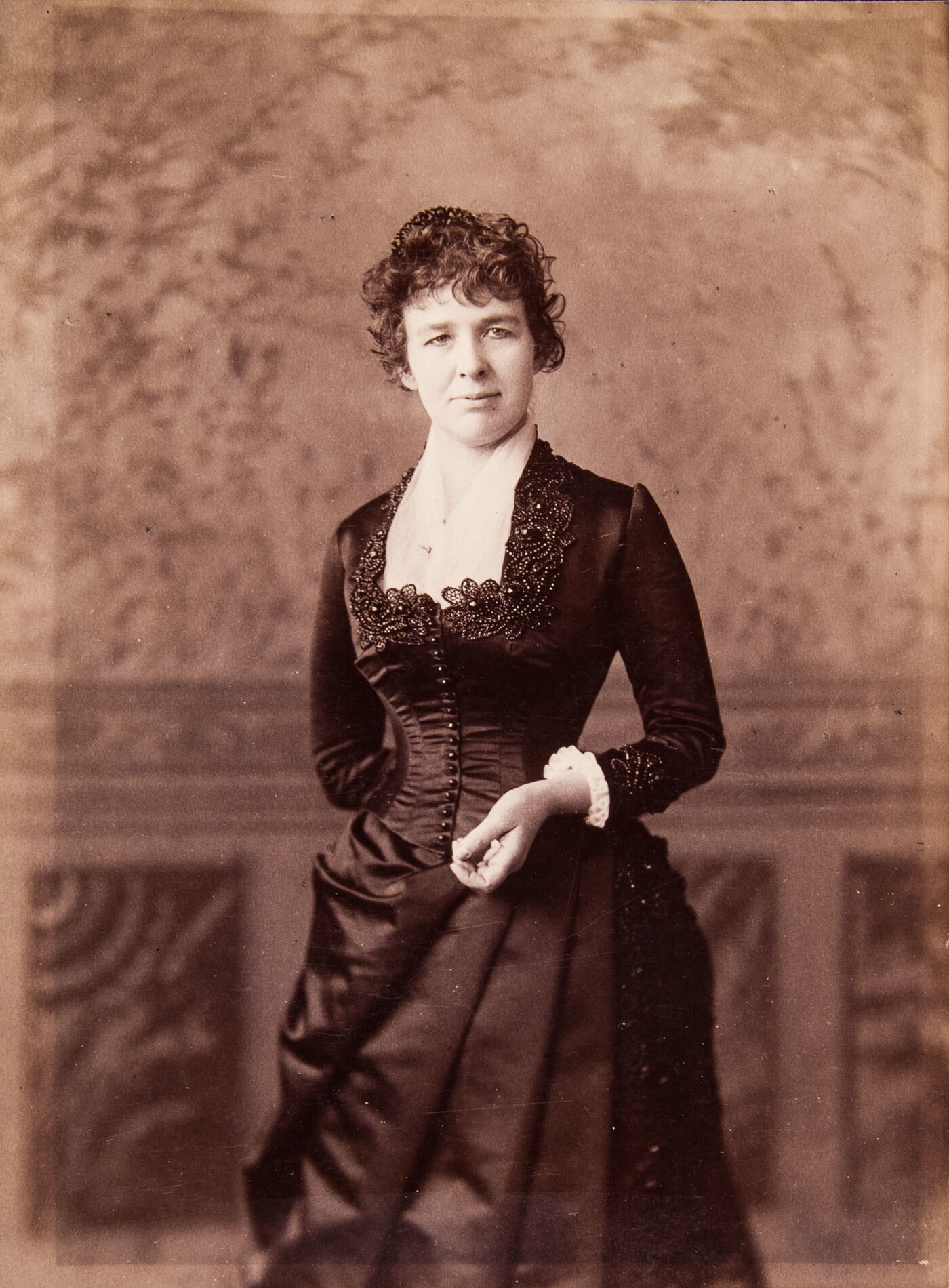 A black-and-white studio portrait photograph of a White woman in a formal, fitted dress standing in front of a backdrop.