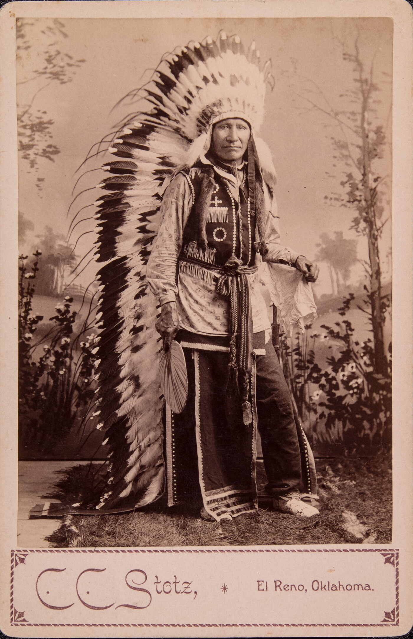A sepia-toned studio portrait photograph of an Indigenous man in full traditional regalia, including a long, feathered headdress.