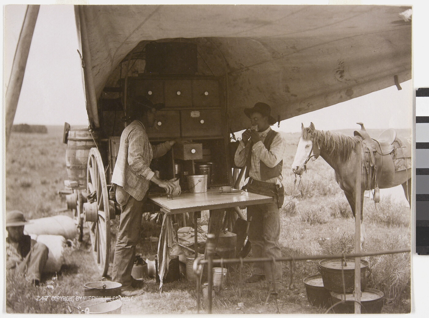 A sepia-toned photograph of two men eating and drinking from a chuckwagon as a horse stands in the background.