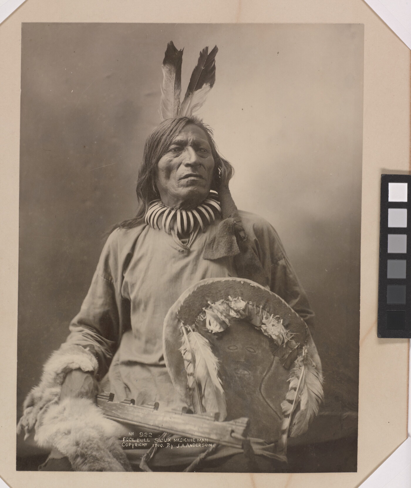 A black-and-white formal portrait photograph of a seated Indigenous man in traditional clothing holding a shield.