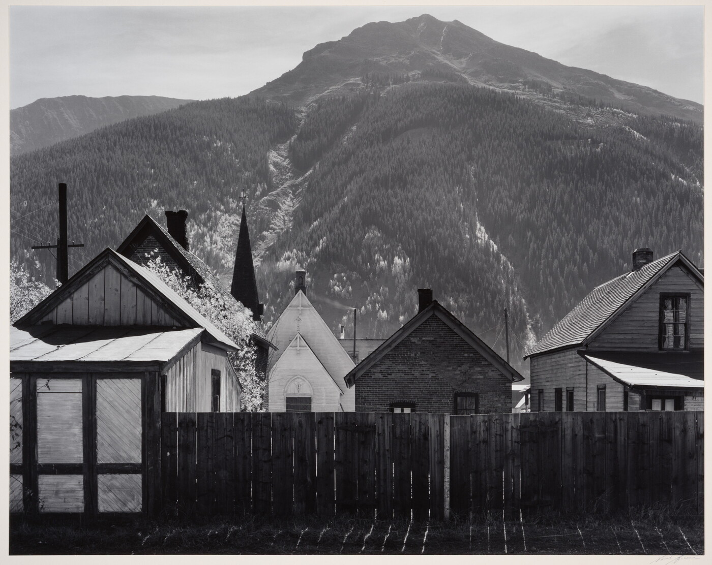 A black-and-white photograph of clapboard houses and a church behind a wood fence at the base of a densely wooded mountain.